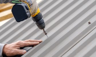 Men's hands in work gloves with a yellow screwdriver screw the roofing sheet to the roof of a country house. Cordless drill. The use of electrical engineering and technology.